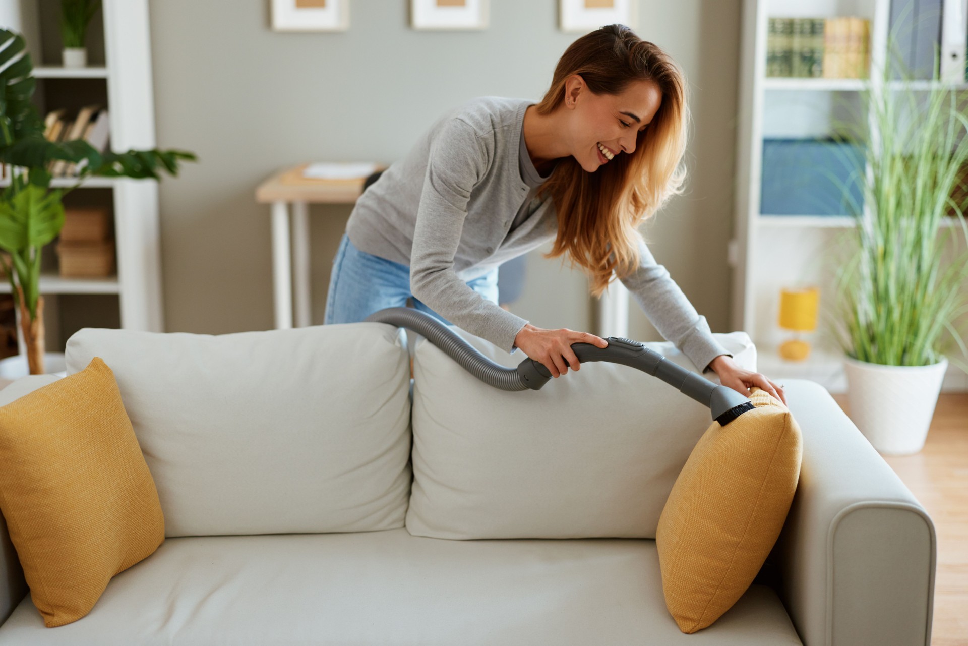 Smiling woman vacuuming sofa in bright living room enjoying housework and domestic chores